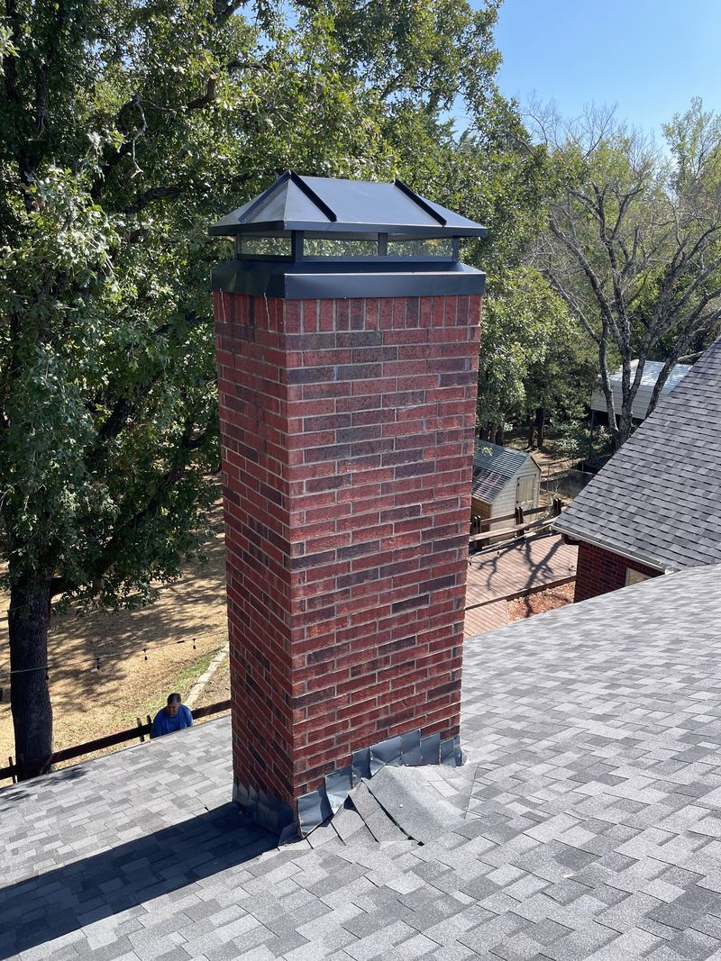 A newly fitted black chimney cap on a red brick chimney above a shingle roof