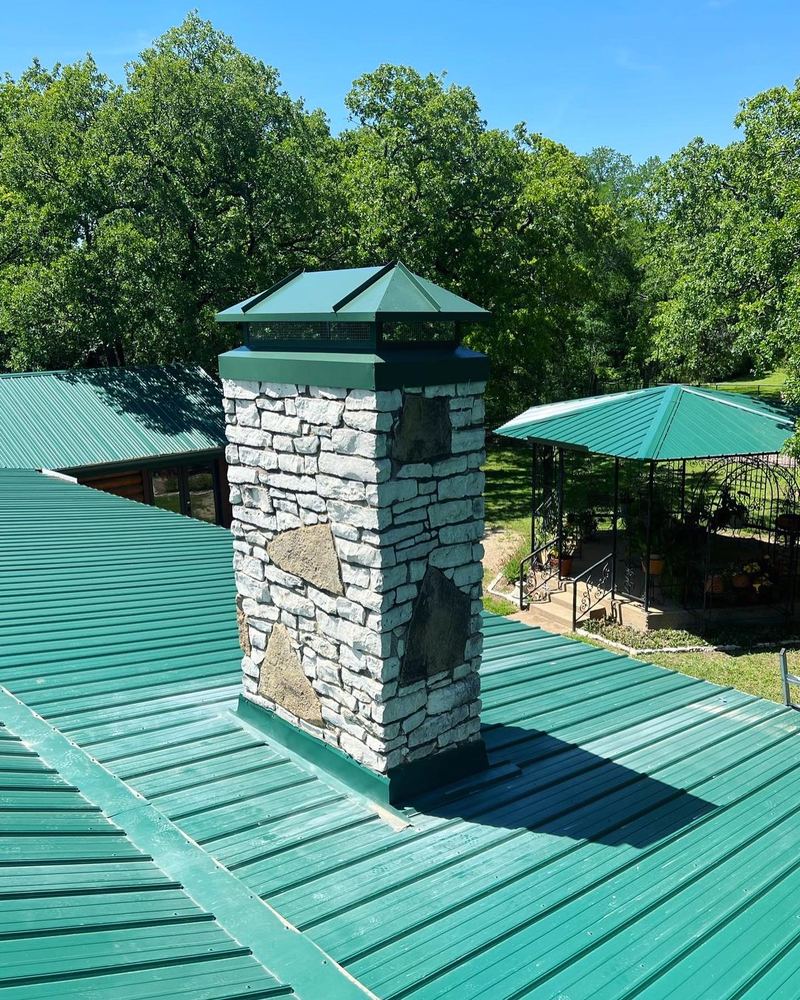A stone chimney with a new cap rising from a green standing seam metal roof